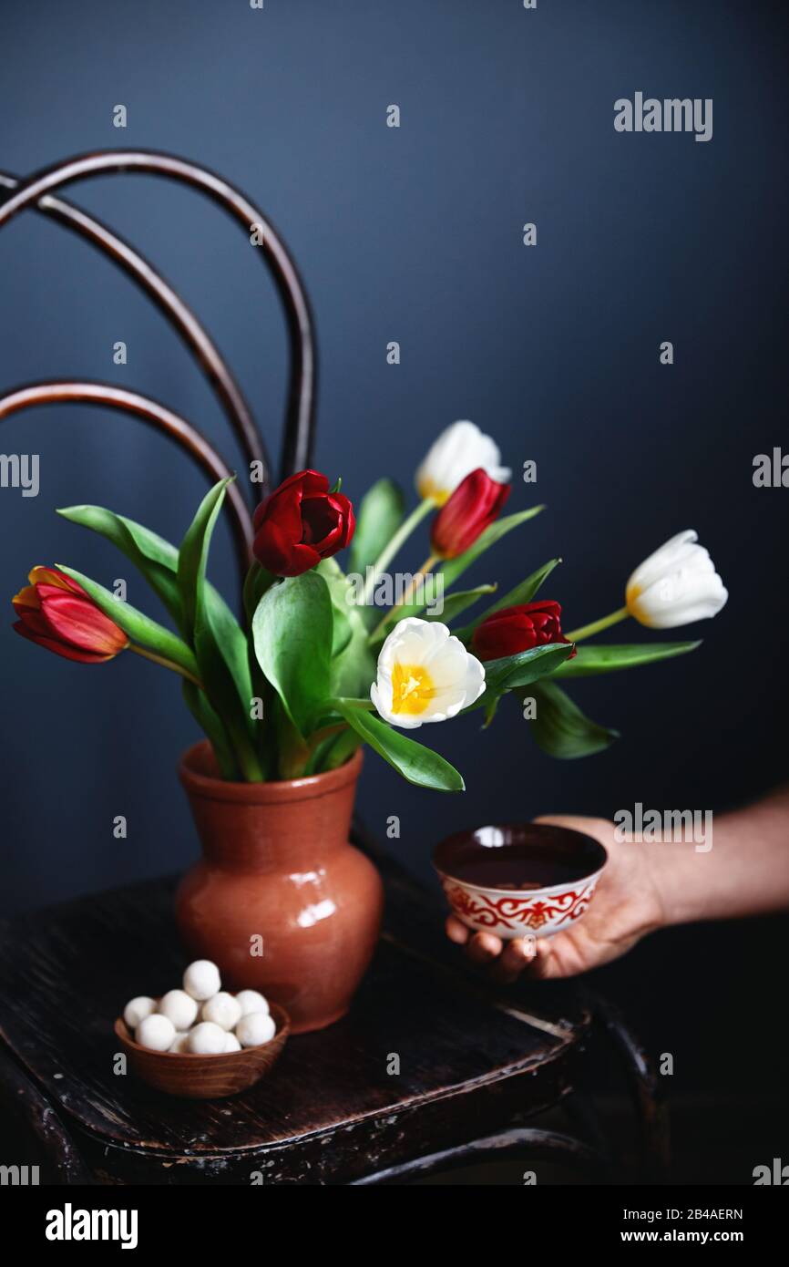 L'homme tenant un bol ethnique avec du thé près du magnifique bouquet de tulipes et des boulettes de kurt sèchent du fromage sur une chaise en bois vintage au festival de Nauryz Banque D'Images