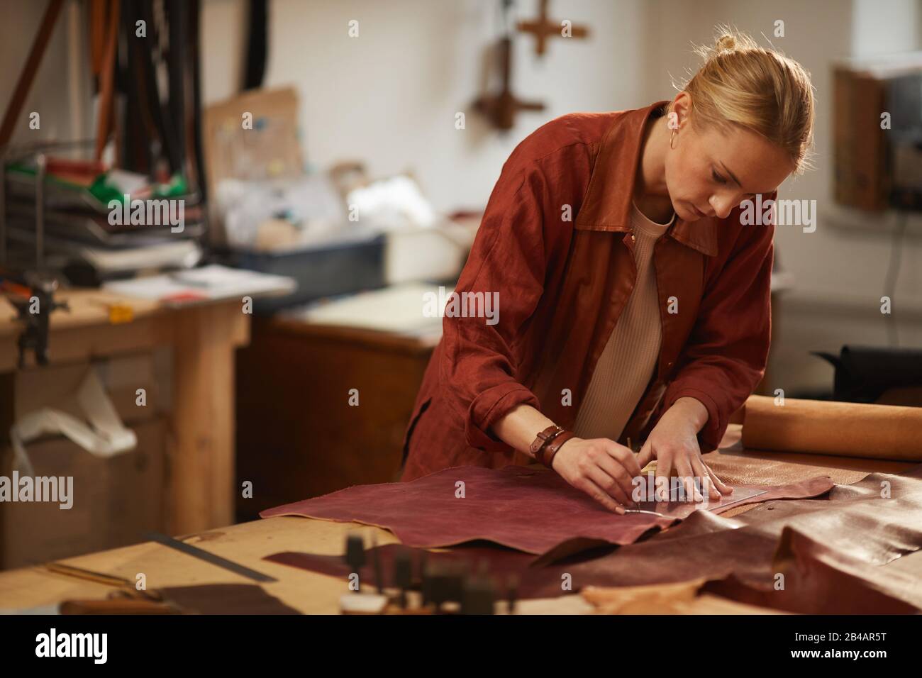 Portrait moyen horizontal de la jeune femme concentrée debout à la table coupant un morceau de cuir Banque D'Images