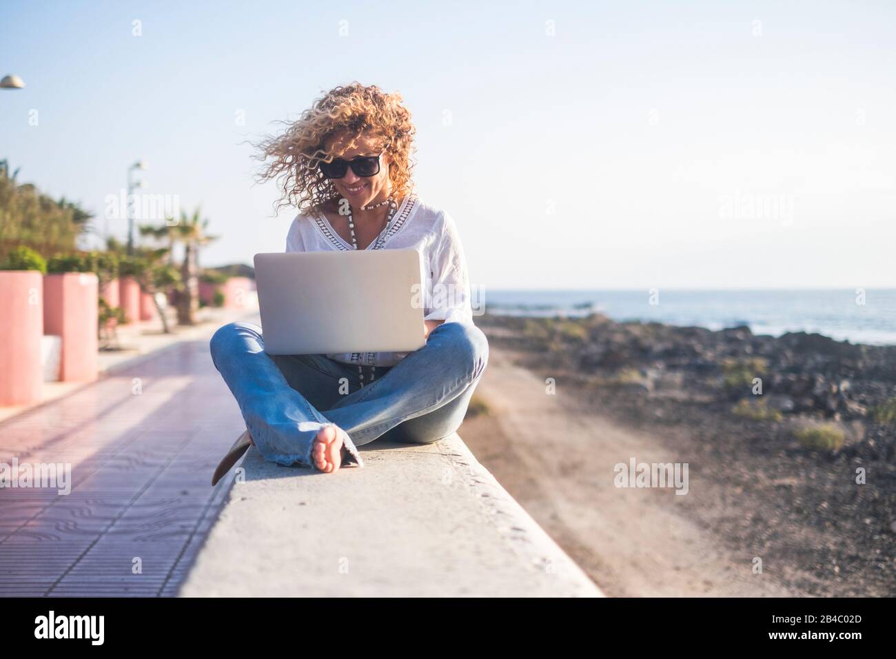 Gai belle blonde curly personnes jeune femme adulte assis en plein air avec la mer et le ciel en arrière-plan travailler avec tablette ordinateur Internet connecté - libre de concept de bureau Banque D'Images