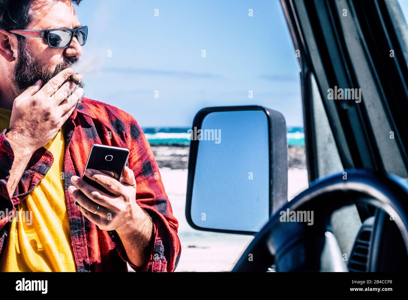 Beau homme debout avec cigare utiliser le téléphone mobile à l'extérieur de sa voiture noire - plage et océan en arrière-plan pour Voyage et aventure style de vie concept - les gens en plein air activité de loisirs Banque D'Images