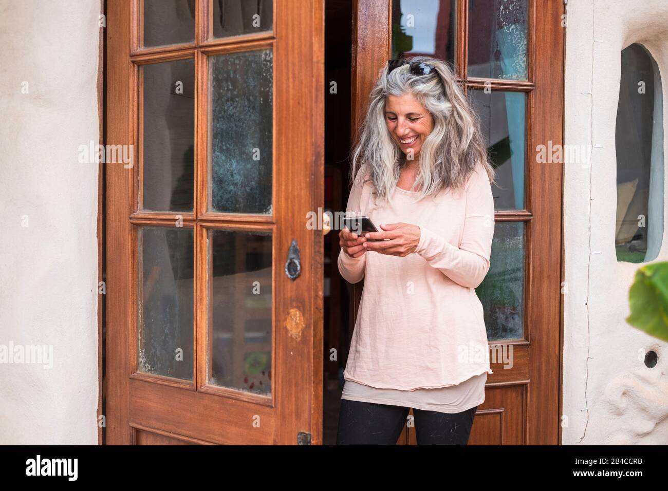 Heureuse et joyeuse femme alternative adulte utilisant la technologie à l'extérieur de la maison dans le jardin - les cheveux gris longs et sourit en utilisant un téléphone mobile hors de la porte Banque D'Images