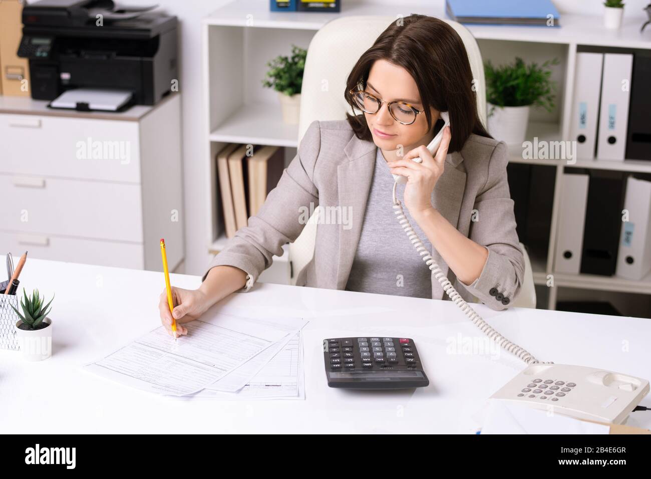 Jolie jeune femme d'affaires élégante assise au bureau, consultant le client au téléphone et prenant des notes dans le document Banque D'Images