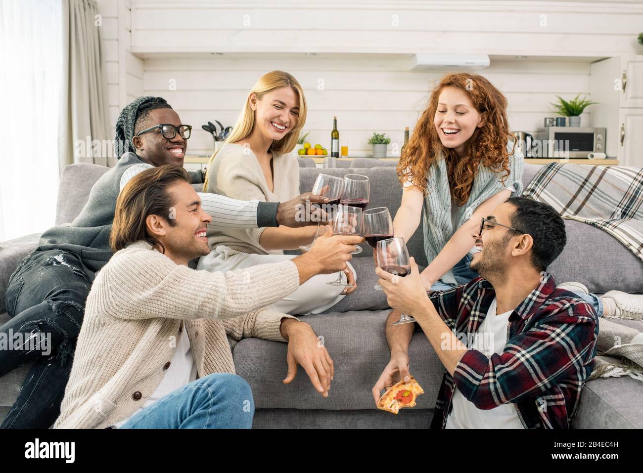 De jeunes amis multiculturels joyeux s'inclinent avec des verres de vin rouge tout en faisant du toast par canapé lors de la fête à la maison Banque D'Images