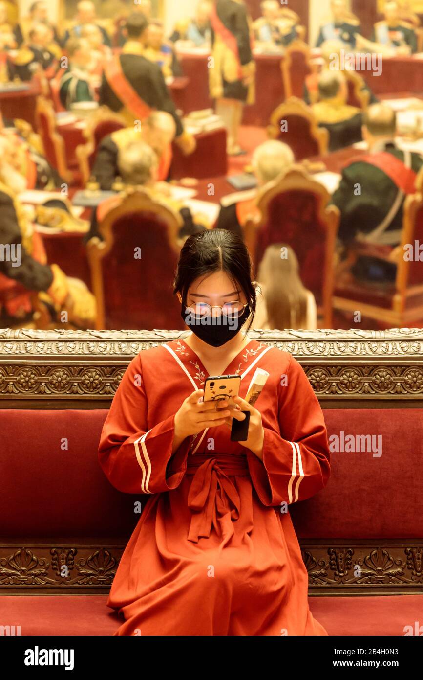 Saint-Pétersbourg, Russie - janv.2020: Jeune femme chinoise en vêtements nationaux portant un masque chirurgical assis dans le musée, à l'aide d'un smartphone. Nouveau type coronav Banque D'Images