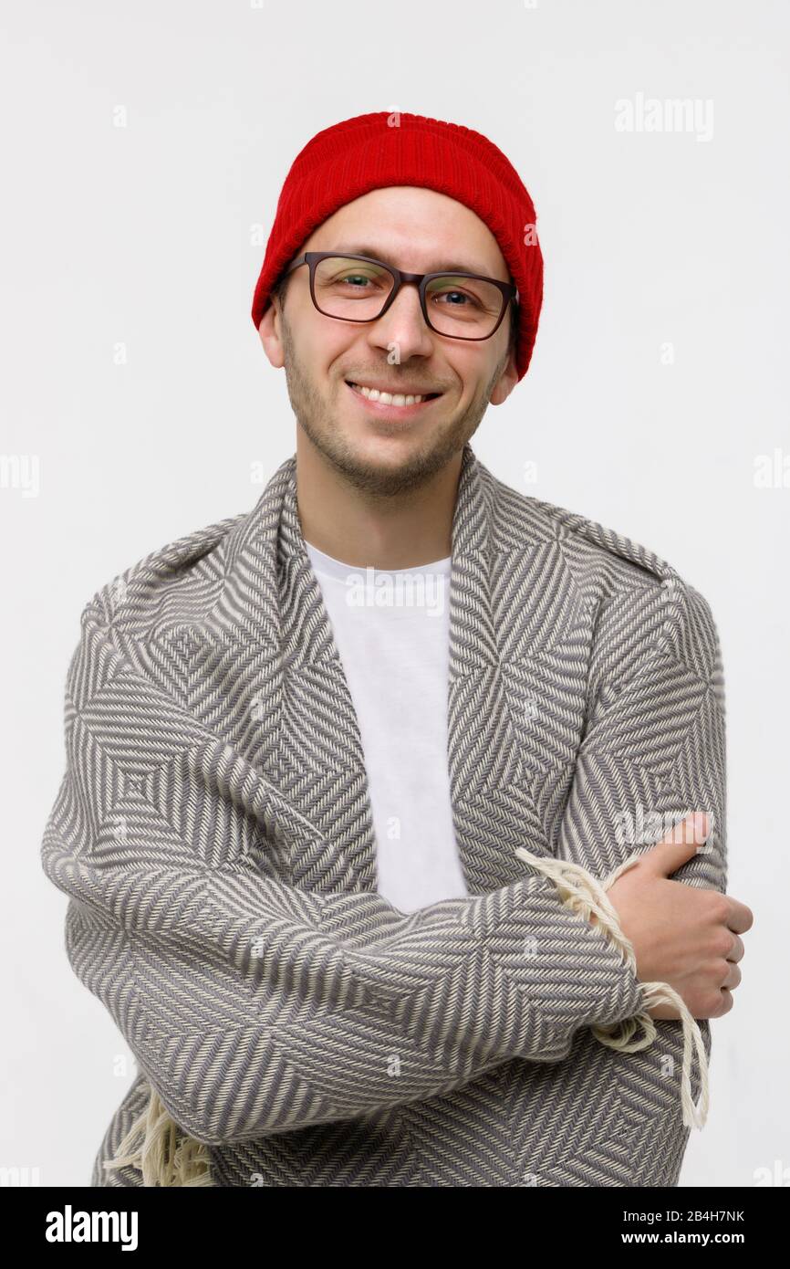 Studio portrait d'un jeune homme plein de gaieté et de style hipster avec des poils légers sur le visage souriant à l'appareil photo, portant des lunettes, un bonnet rouge et enveloppé dans un plat, un Banque D'Images
