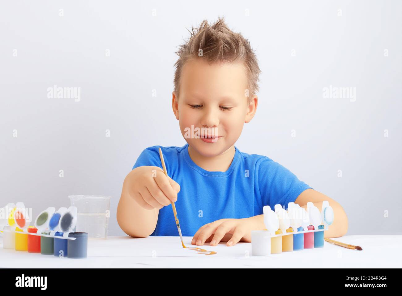 Un jeune garçon joyeux et joyeux aux cheveux bousieux tient une brosse dans sa main et dessine avec de la peinture dorée. Concept d'éducation à l'école ou à la maternelle. Banque D'Images