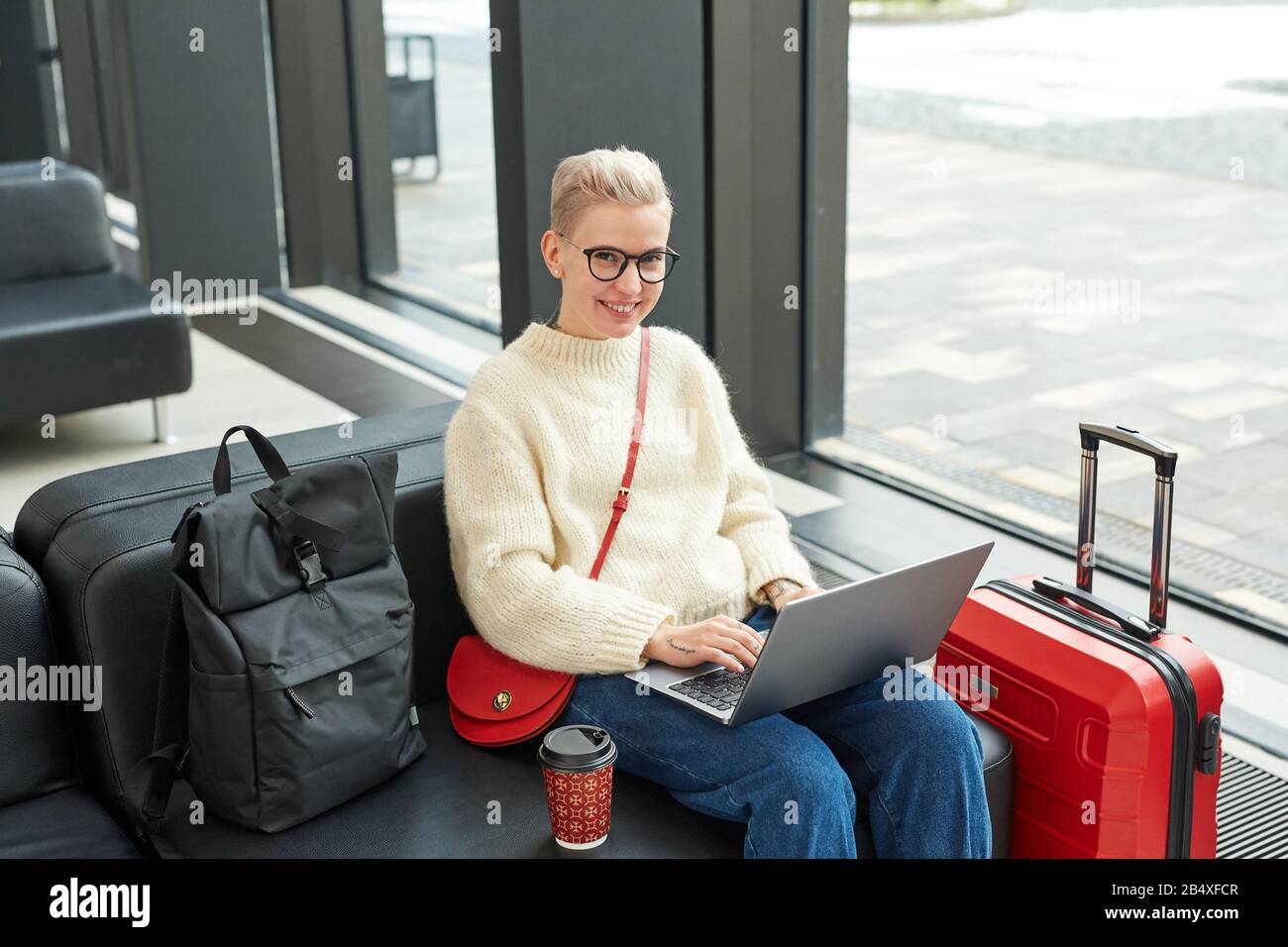 Photo horizontale en grand angle d'une femme blonde élégante portant des lunettes assis dans le salon de départ à l'aide de son ordinateur portable regardant l'appareil photo Banque D'Images