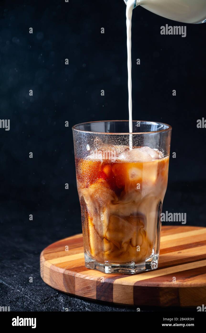 Le Lait Est Versé Dans Du Café Glacé Dans Le Grand Verre Sur Fond Sombre. Boisson D'Été Rafraîchissante Concept. Banque D'Images