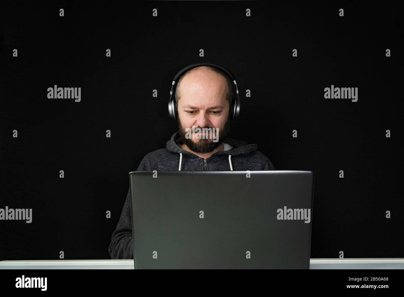 Homme caucasien dans le hoodie et les écouteurs assis derrière l'ordinateur portable et de travail, fond noir, concept de bureau à la maison Banque D'Images
