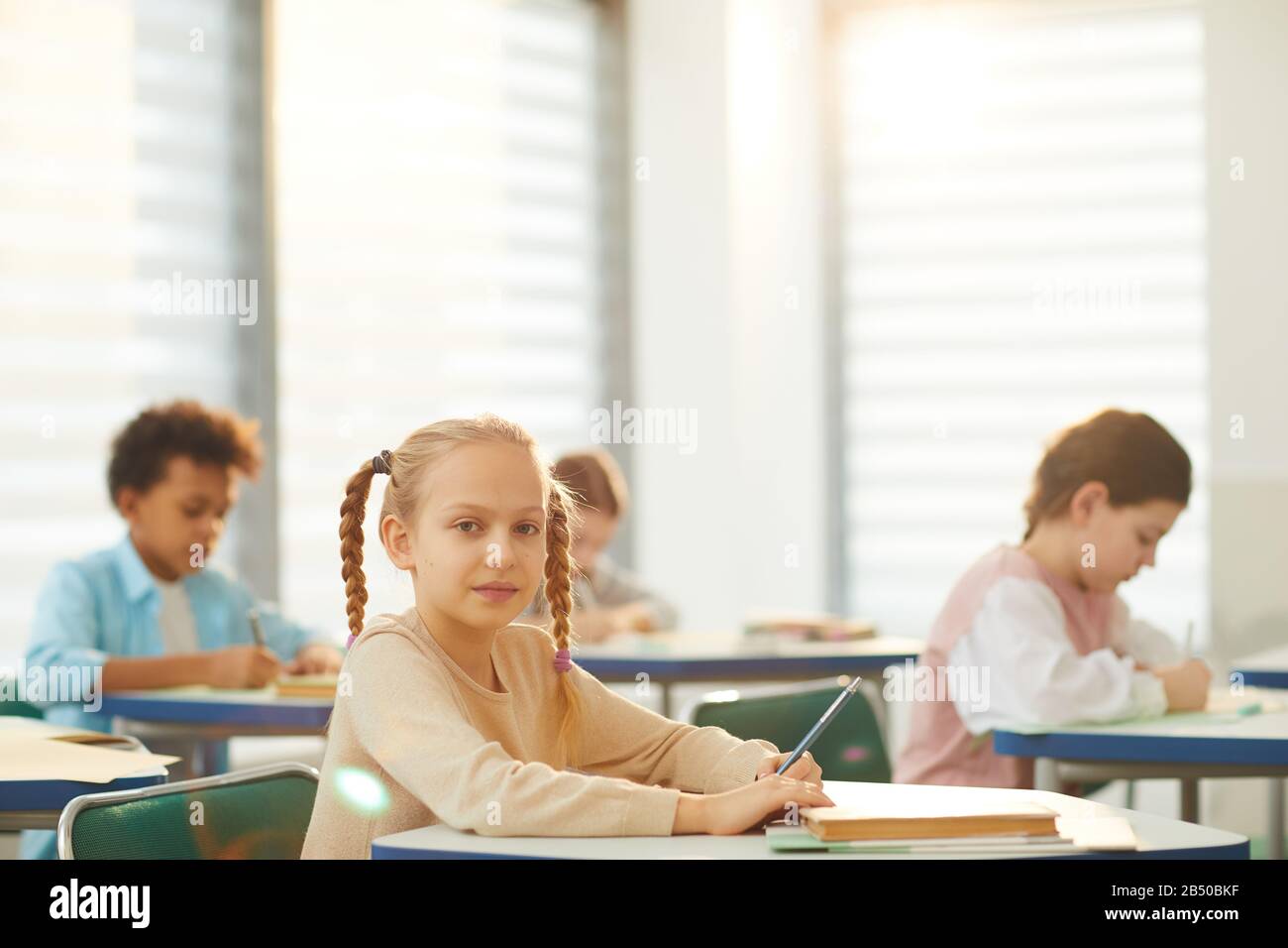 Portrait horizontal de la petite fille caucasienne avec des cheveux blonds assis à la table de l'école tenant crayon regardant l'appareil photo, espace de copie Banque D'Images