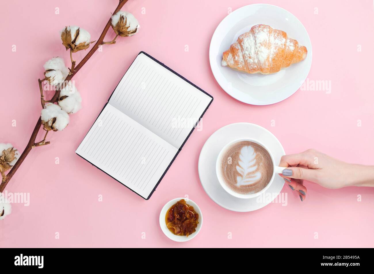 Les mains retiennent le café avec de l'art latte et un cahier avec croissant, confiture et branche en coton Banque D'Images