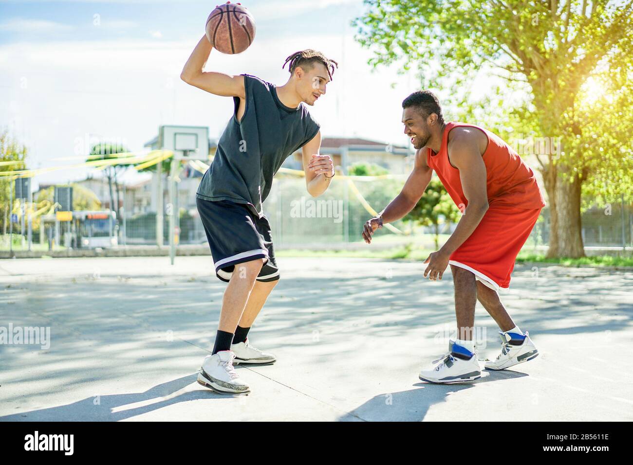 Deux amis joyeux et multiraciaux qui jouent au basket-ball en plein air dans le cadre d'un concours urbain avec contre-jour : les jeunes qui s'amusent à faire du sport pour une bonne santé Banque D'Images