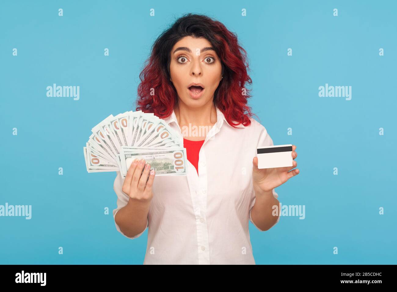 WOW, prêt bancaire pour les achats. Portrait d'une femme hipster surprise avec des cheveux rouges fantaisie tenant la carte de crédit, des billets de dollar et regardant avec étonnement choqué Banque D'Images