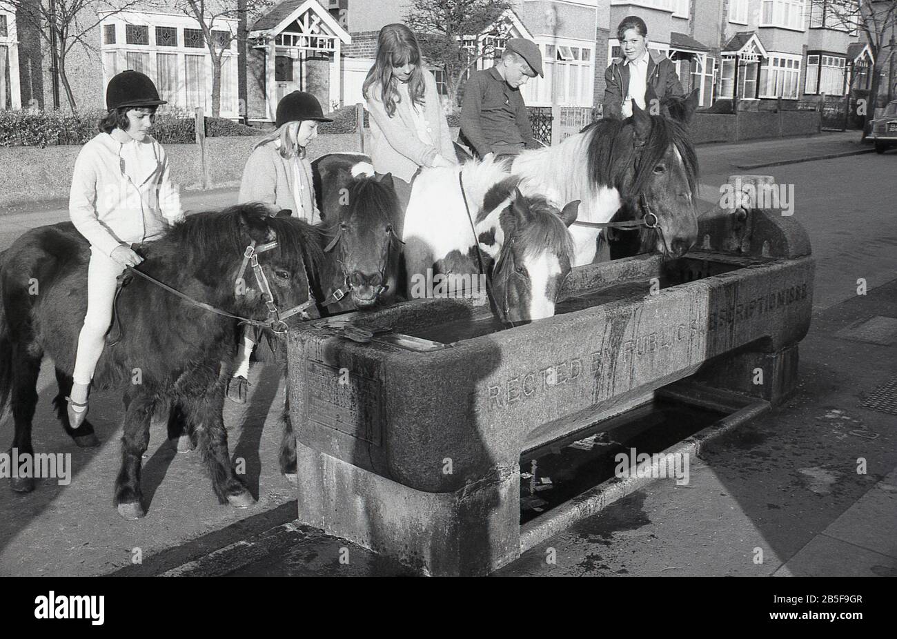 Les années 1970, historiques, dans une rue suburbaine, les jeunes sur les chevaux et les poneys s'arrêtent dans une ancienne auge d'eau en pierre - construite en 1888 - pour donner à leurs animaux rafraîchissement, Londres du Sud, Angleterre, pendant un été chaud particulier. Au cours de la décennie, certains étés étaient extrêmement chauds. Banque D'Images