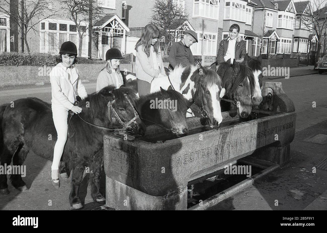 Les années 1970, historiques, dans une rue suburbaine, les jeunes sur les chevaux et les poneys s'arrêtent dans une ancienne auge d'eau en pierre - érigée en 1888 - pour donner à leurs animaux rafraîchissement, Londres du Sud, Angleterre, pendant un été chaud particulier. Au cours de la décennie, certains étés étaient extrêmement chauds. Banque D'Images