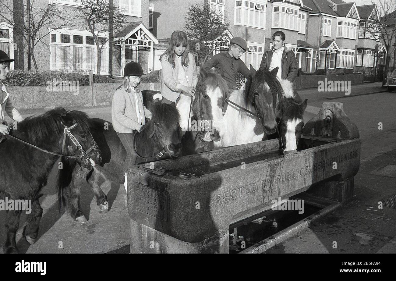 Les années 1970, historiques, dans une rue suburbaine, les jeunes sur les chevaux et les poneys s'arrêtent dans une ancienne auge d'eau en pierre - érigée en 1888 - pour donner à leurs animaux rafraîchissement, Londres du Sud, Angleterre, pendant un été chaud particulier. Au cours de la décennie, certains étés étaient extrêmement chauds. Banque D'Images