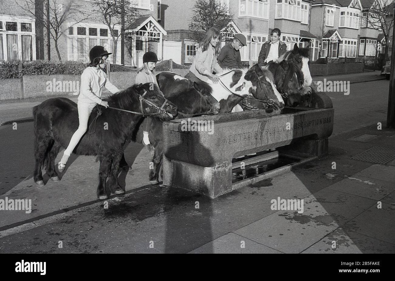 Les années 1970, historiques, dans une rue suburbaine, les jeunes sur les chevaux et les poneys s'arrêtent dans une ancienne auge d'eau en pierre - érigée en 1888 - pour donner à leurs animaux rafraîchissement, Londres du Sud, Angleterre, pendant un été chaud particulier. Au cours de la décennie, certains étés étaient extrêmement chauds. Banque D'Images