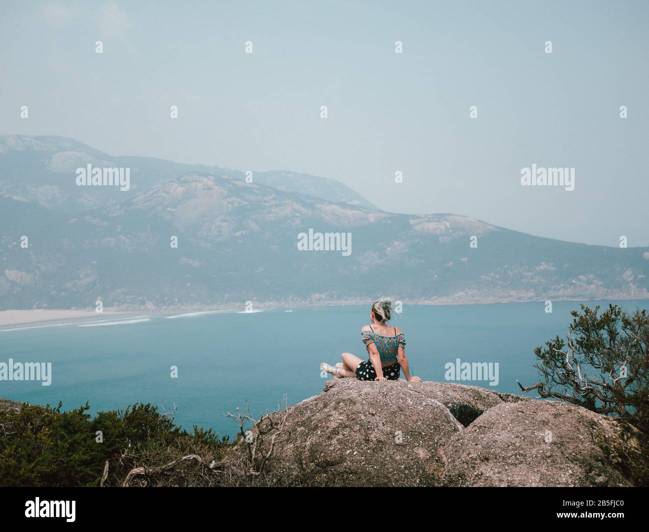 Une jeune femme assise au bord d'un rocher au sommet d'une montagne qui donne sur un magnifique bleu océan Banque D'Images