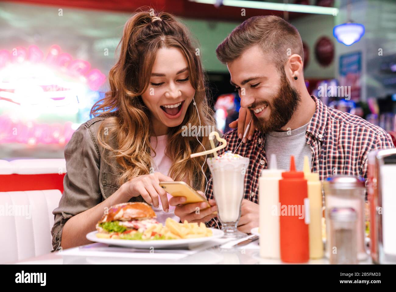 L'image d'un jeune couple joyeux et joyeux s'assoit dans un café de cuisine de rue rétro lumineux utilisant la musique d'écoute de téléphone mobile avec écouteurs. Banque D'Images