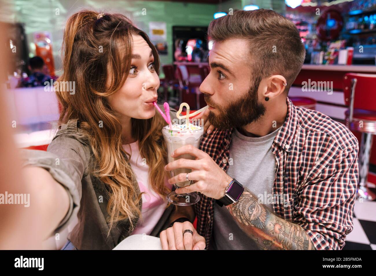 Image d'un drôle de jeune couple épris positif émotive dans le café rétro lumineux de la nourriture de rue prendre un selfie par l'appareil photo buvant cocktail. Banque D'Images