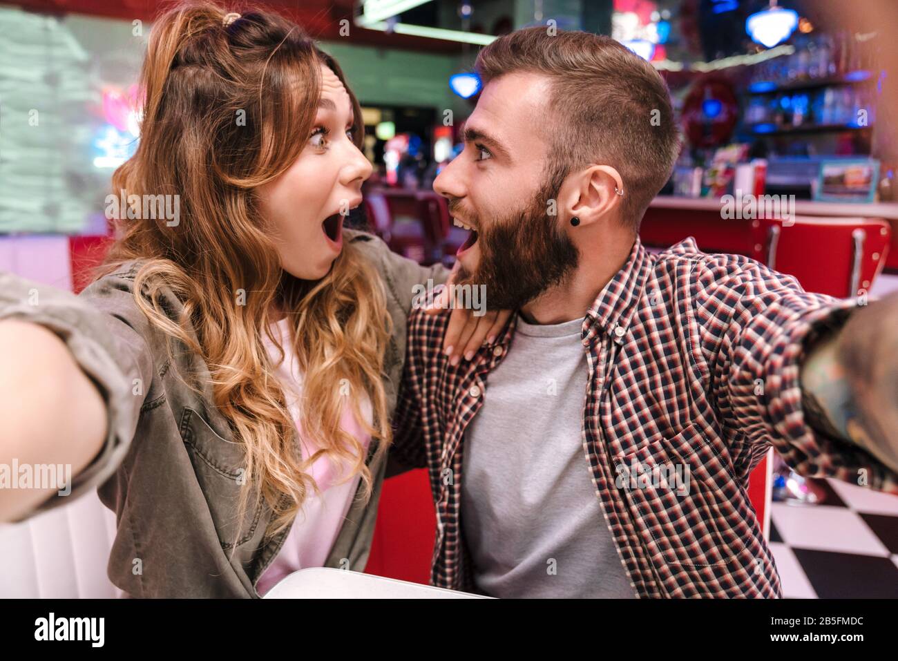 Image d'un joli couple surpris et enchanté de jeunes amoureux dans un café de cuisine de rue rétro lumineux prendre un selfie par appareil photo. Banque D'Images