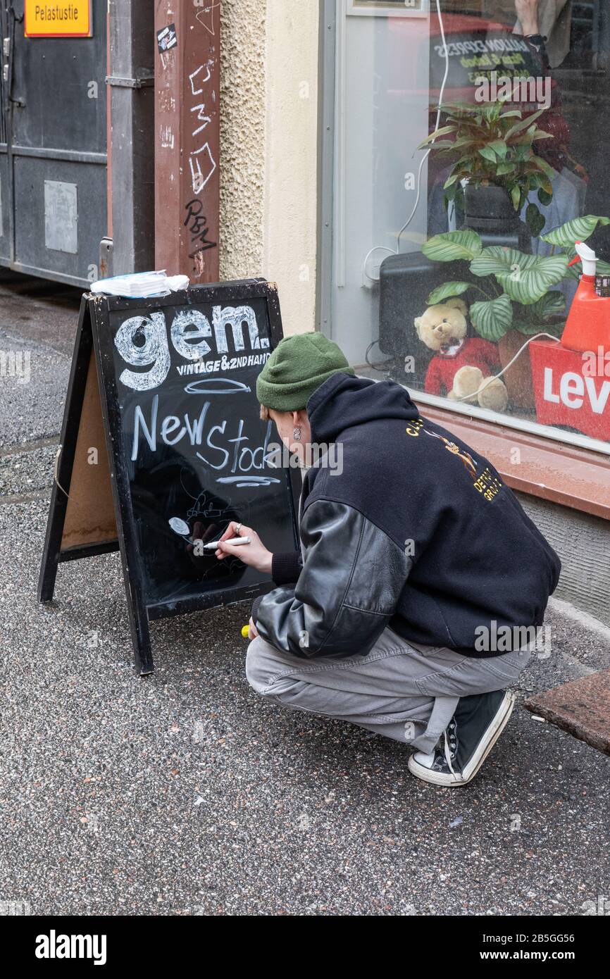Jeune homme écrivant sur un tableau de bord latéral devant Gem vintage et magasin secondaire dans le district de Kallio à Kallio, en Finlande Banque D'Images