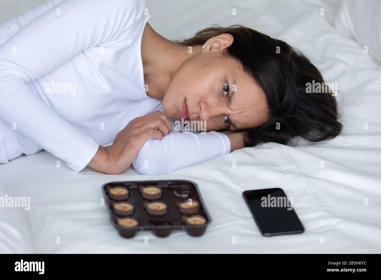 Fille déprimée posée dans le lit avec des bonbons et smartphone Banque D'Images