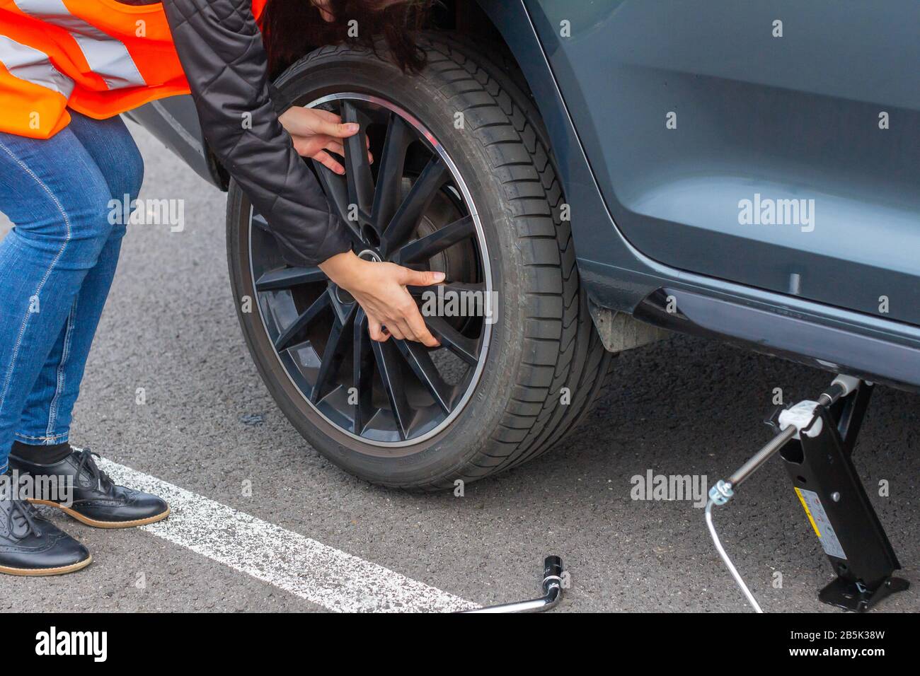 Une femme dans un gilet de sécurité orange change une roue dans une voiture cassée sur la route. Approcher les mains des femmes de la roue d'un véhicule, en essayant de changer un plat Banque D'Images