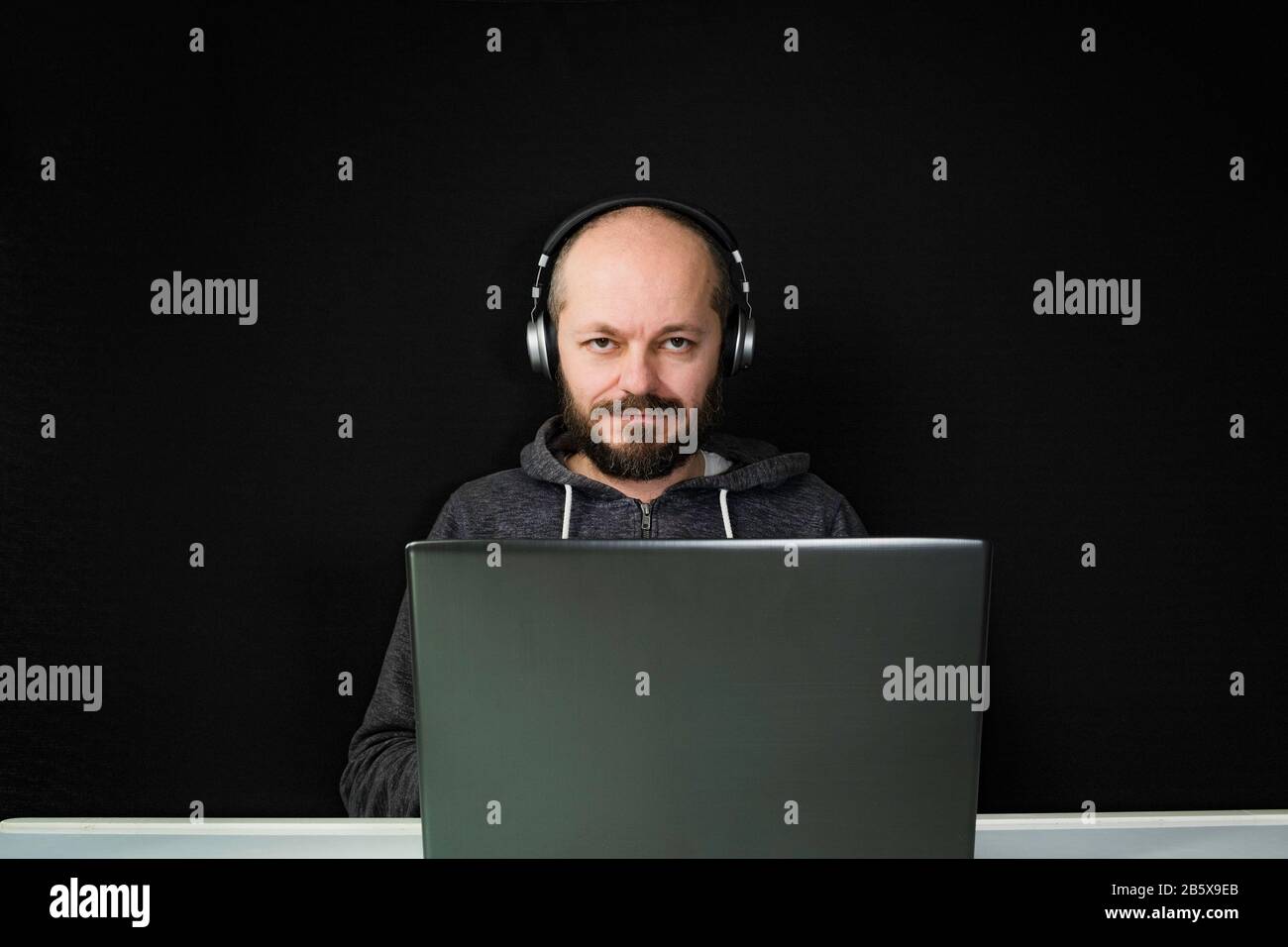 Homme caucasien dans le hoodie et les écouteurs assis derrière l'ordinateur portable et de travail, fond noir, concept de bureau à la maison Banque D'Images