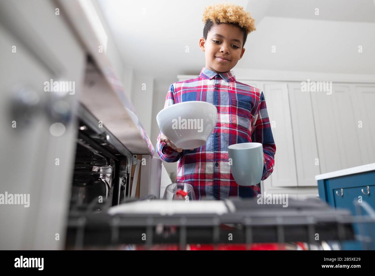 Garçon Aider Avec Les Corvées À La Maison En Empiétant Vaisselle Dans Le Lave-Vaisselle Banque D'Images