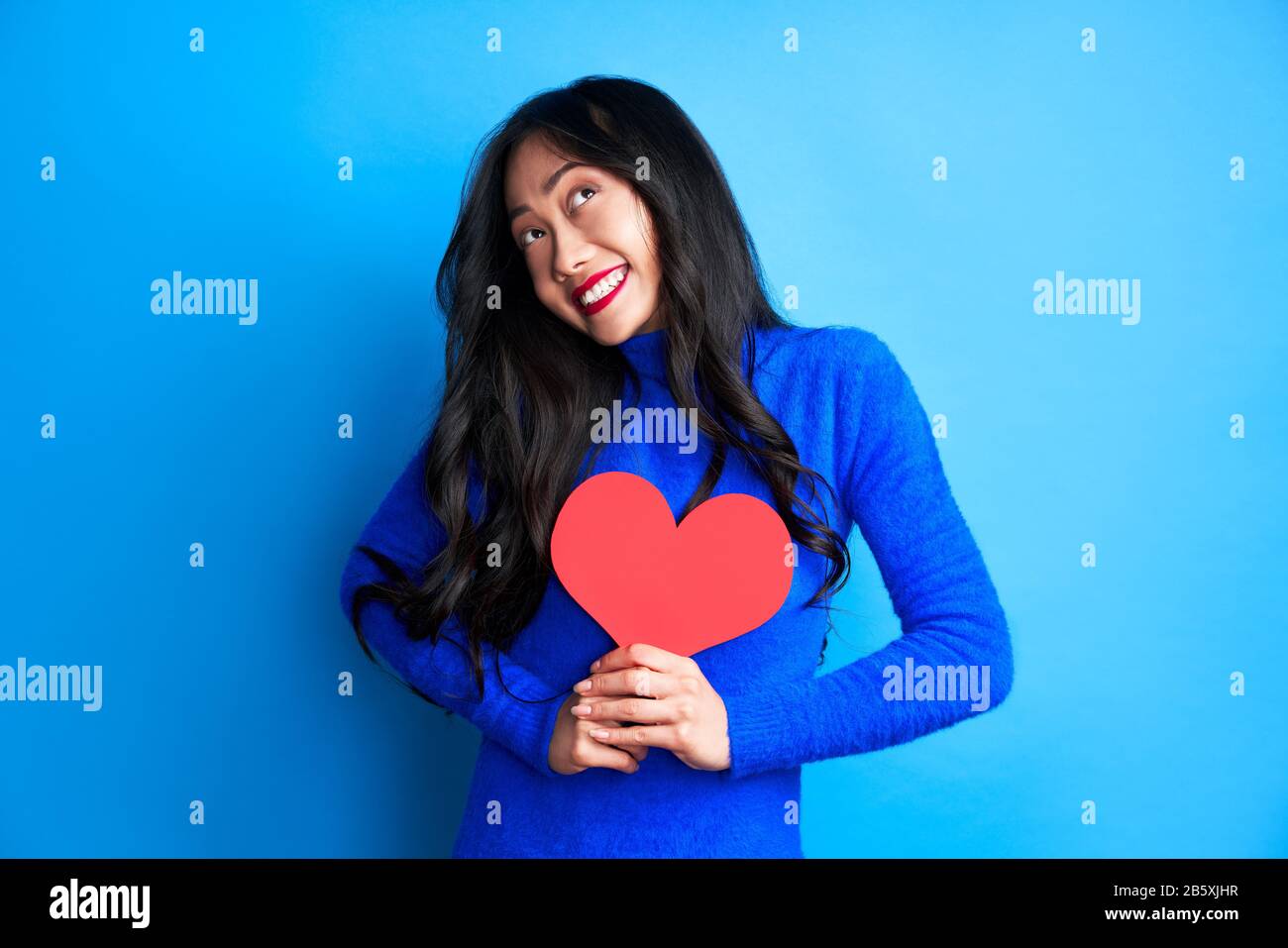 Une femme souriante et heureuse qui tient un grand cœur en papier dans ses mains, regarde isolée sur fond bleu. Concept d'amour Banque D'Images
