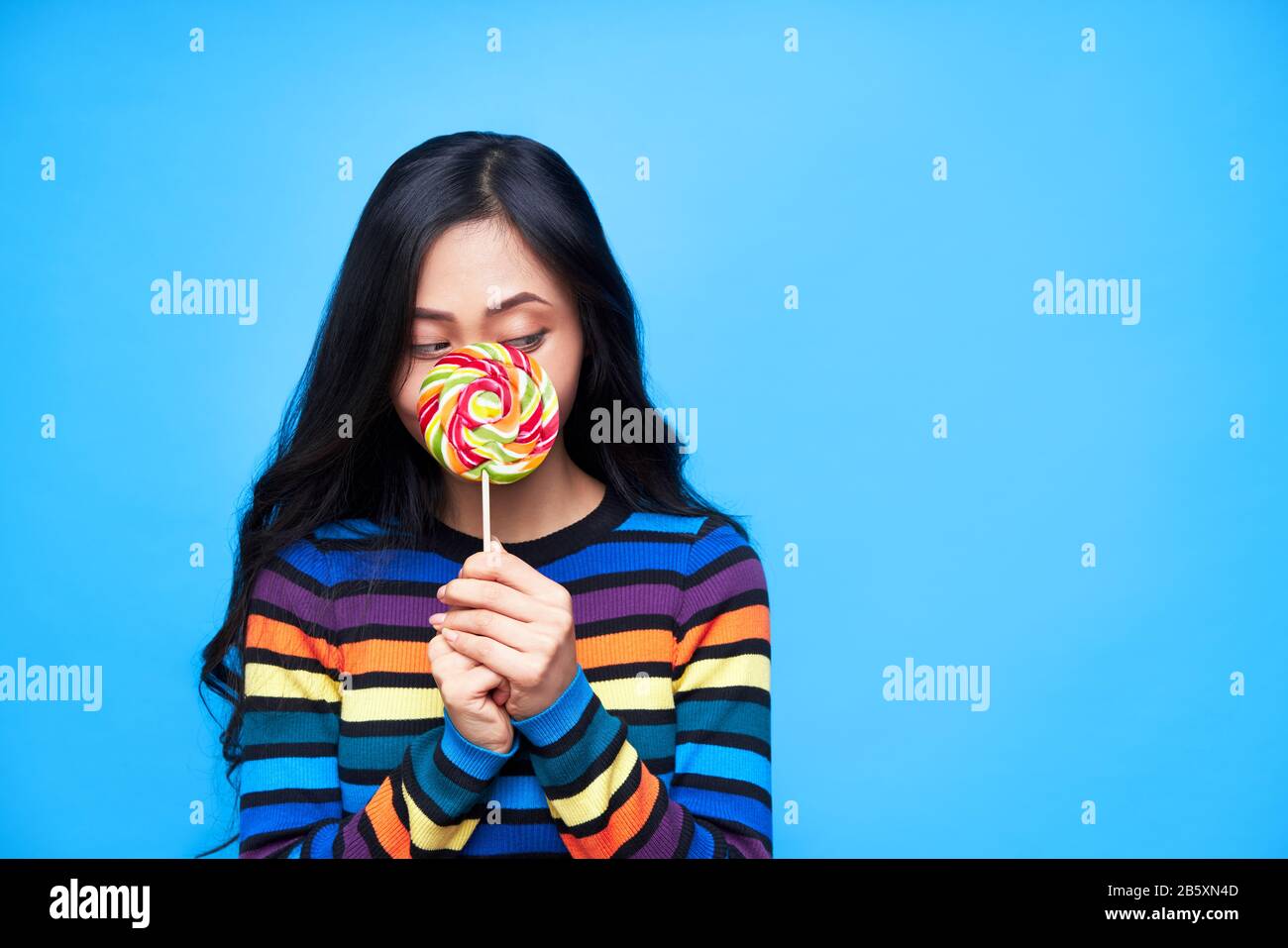 La jeune femme cache son visage derrière un lollipop coloré isolé sur fond bleu. Cocept d'émotion, nourriture douce Banque D'Images