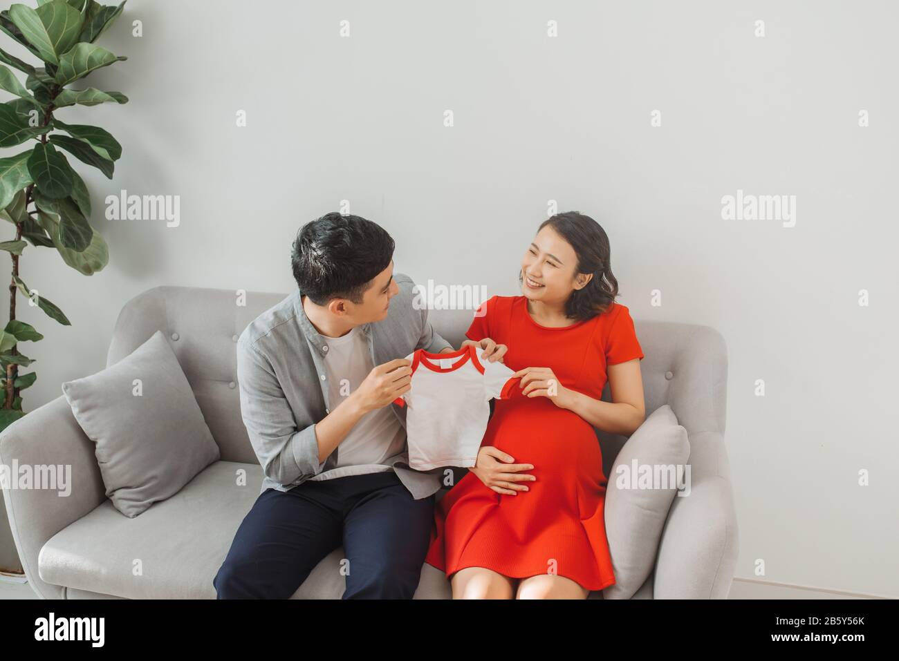 Un jeune mari et une femme enceinte présentent leur t-shirt bébé lorsqu'ils sont assis sur un canapé. Banque D'Images