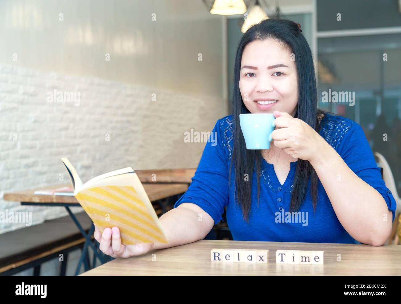 Les femmes d'origine ethnique asiatique détiennent une tasse de céramique pour boire un café chaud ou du thé et lisent un livre dans un café. Sur une table en bois, il y a une lettre en bois Banque D'Images