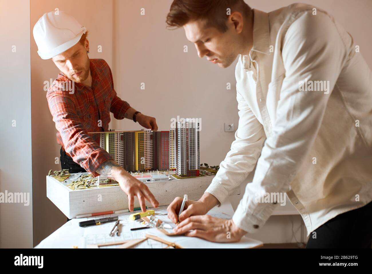 jeune gars barbu dans l'helment pointant vers le plan directeur, donnant des conseils à son collègue qui dessine une maison sur le papier dans le bureau avec le moderne moi Banque D'Images