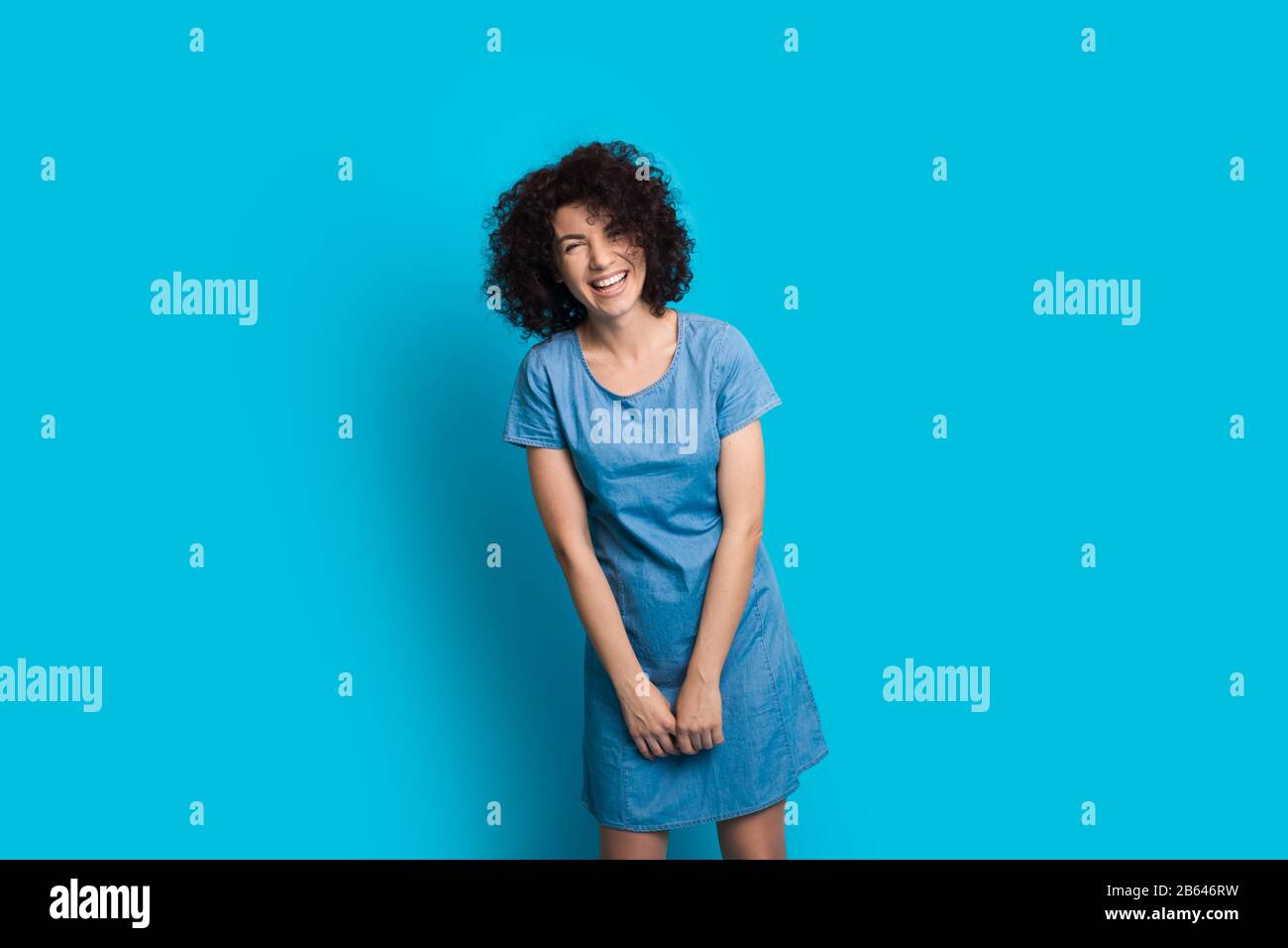 Une femme caucasienne aux cheveux bouclés se posant dans une jolie robe sur un fond bleu tout en souriant chaleureusement Banque D'Images