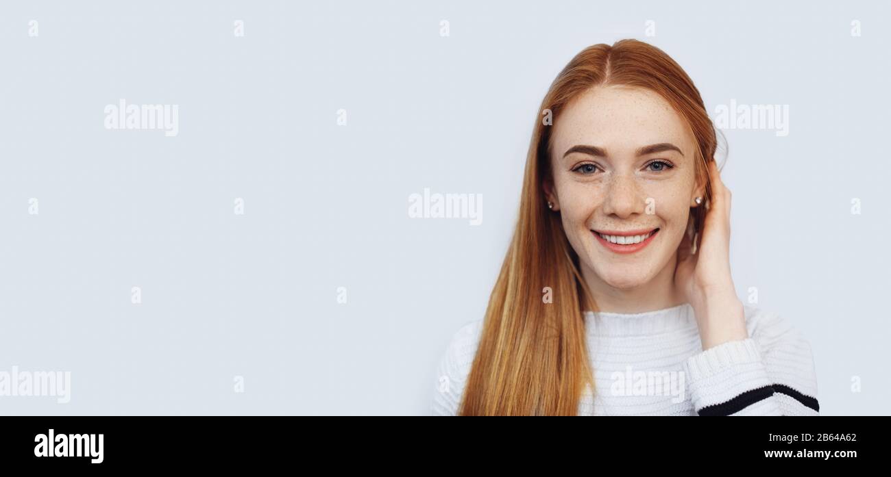 Une charmante fille aux cheveux rouges avec des taches de rousseur touche ses cheveux tout en regardant l'appareil photo et en souriant sur le mur blanc avec de l'espace libre Banque D'Images