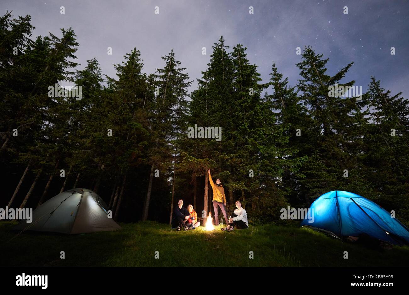 Guy montre à ses amis le ciel étoilé au-dessus de la ville de tente. Les touristes sont assis au coin du feu sur fond de forêt d'épicéa et profitant du ciel du soir. Loisirs en plein air Banque D'Images