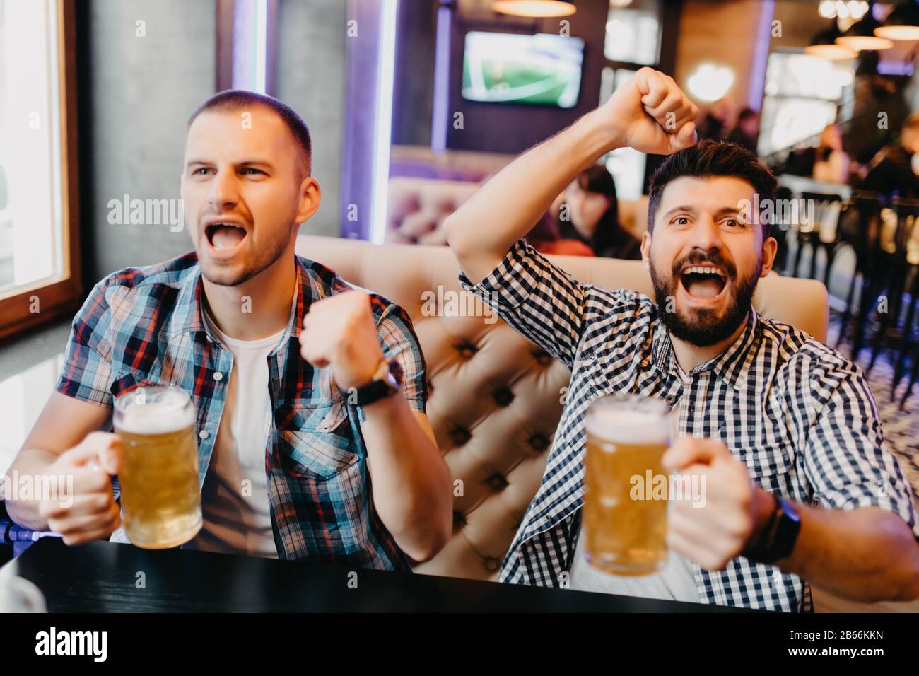 Fans deux jeunes hommes en chemise de plaid au bar ont levé les mains avec des verres de bière et criant, émotions de jeu passionnant à la télévision Banque D'Images