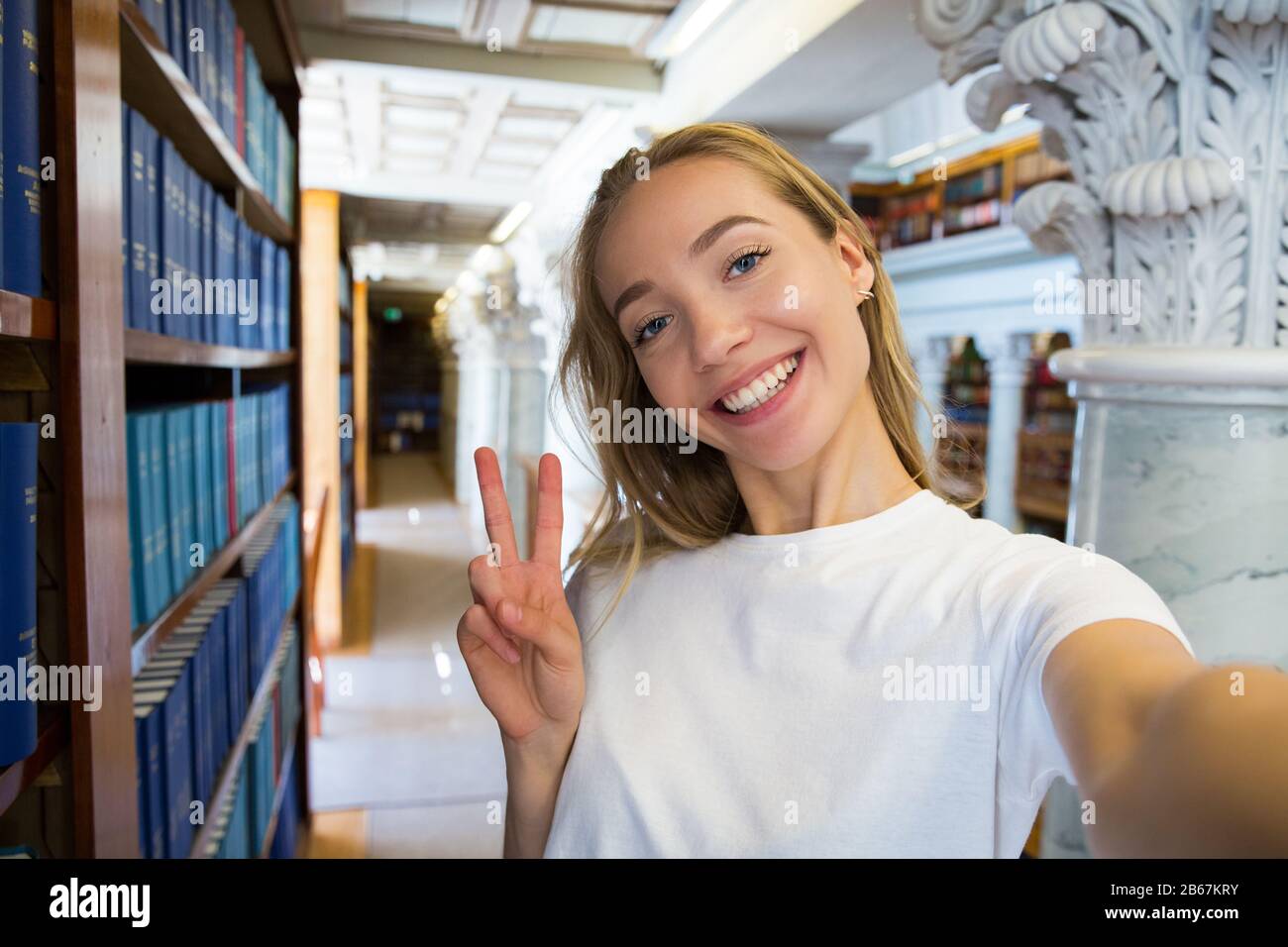 Jeune fille excitée debout dans la vieille bibliothèque traditionnelle à la bibliothèque, souriant et riant étudiant faisant selfie sur l'appareil photo de téléphone, ayant plaisir. Supérieur Banque D'Images