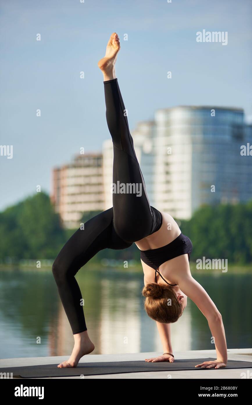 Jeune fille yogi attrayante faisant du yoga, debout dans l'exercice de pont, une pose de roue à pattes, porter des vêtements de sport noirs, arrière-plan résidentiel frais, lac de ville, vue latérale, pleine longueur Banque D'Images