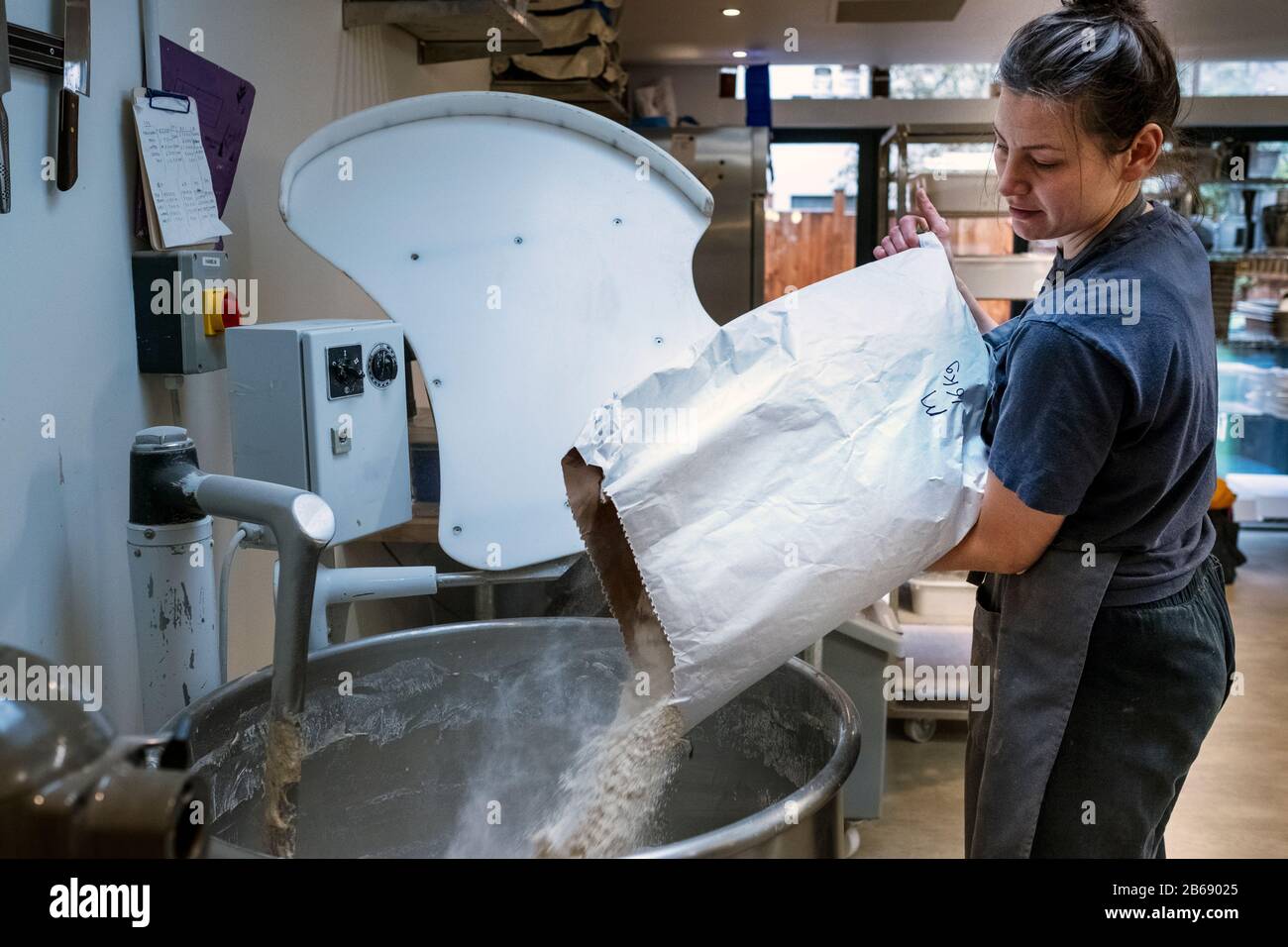 Femme portant un tablier debout dans une boulangerie artisanale, en versant de la farine dans un mélangeur industriel. Banque D'Images