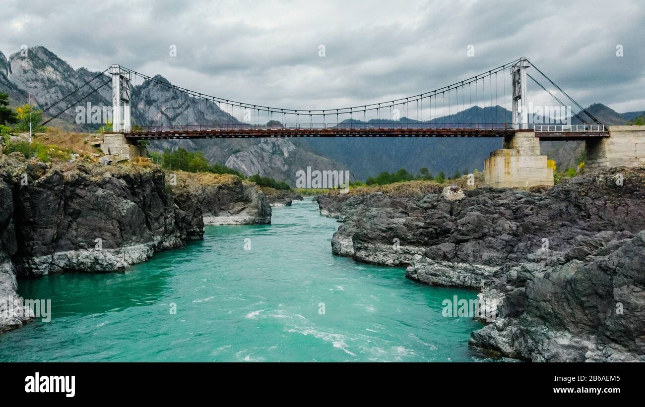 Pont suspendu au-dessus d'une rivière de montagne. Vue depuis la rivière. Rivière turquoise des montagnes, rives rocheuses. Tir drone. Rivière Katun, Altai. Banque D'Images