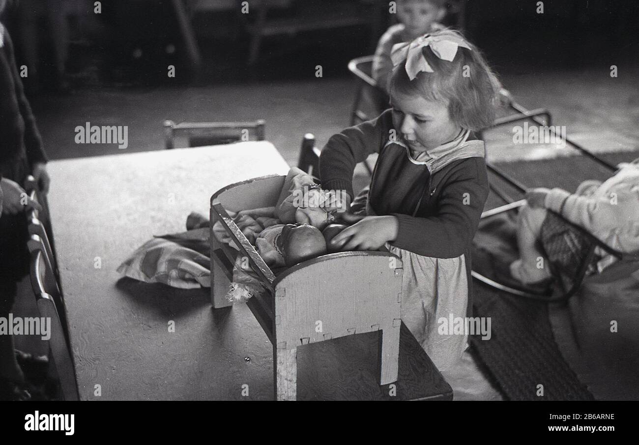 1950, historique, la Grande-Bretagne d'après-guerre et à l'intérieur d'une  chambre dans une pépinière ou une école maternelle, une petite fille jouant  avec des poupées dans un petit lit de jouet en bois.