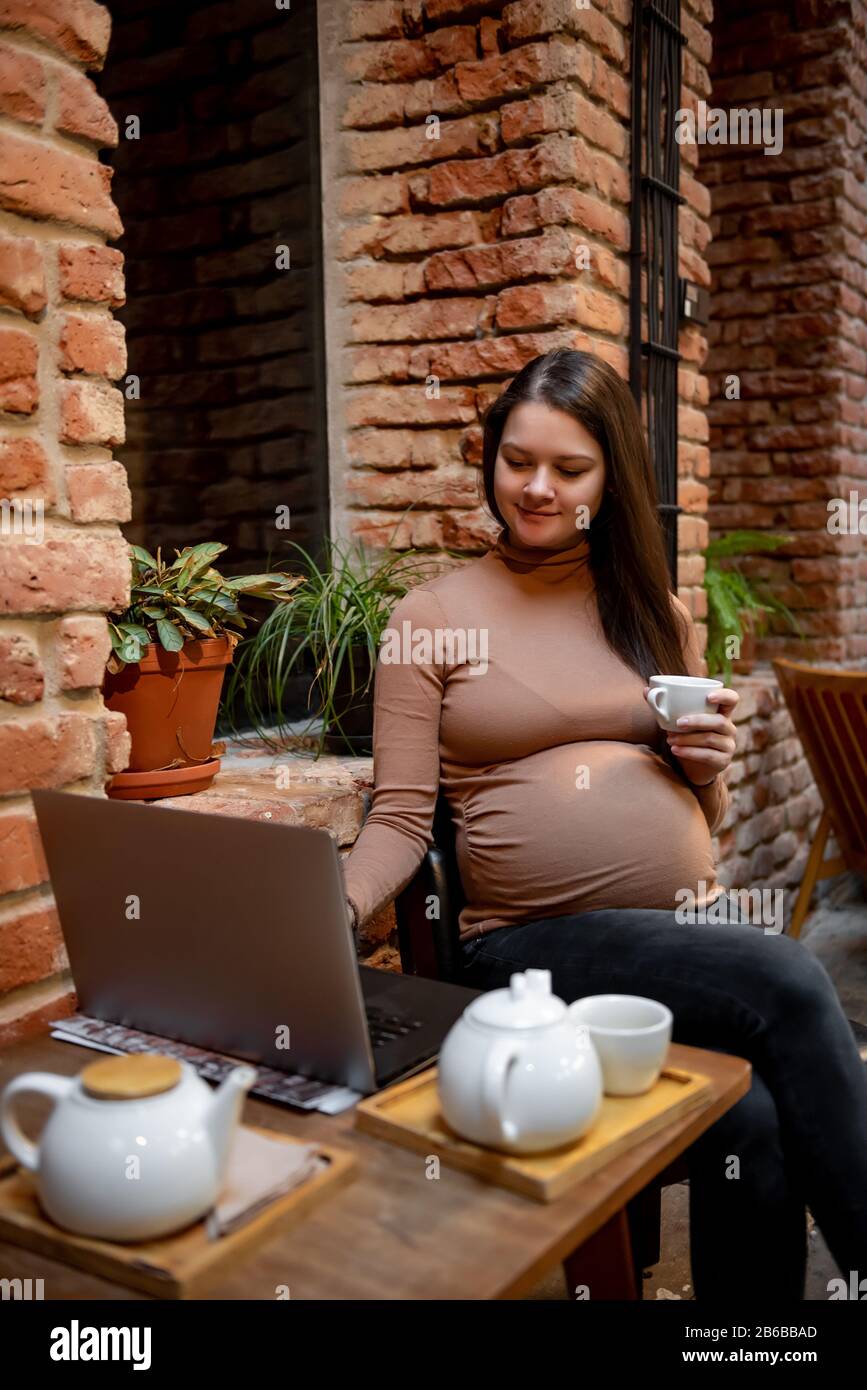 Portrait de jeune femme indépendante prégnanty utilisant un ordinateur portable pour un travail à distance tout en étant assis dans un café moderne avec une tasse de thé Banque D'Images