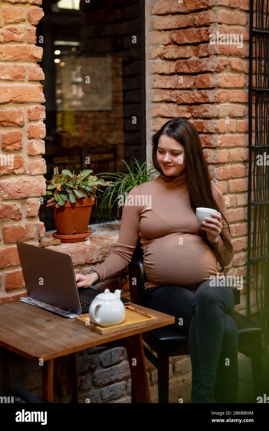 Portrait de jeune femme indépendante prégnanty utilisant un ordinateur portable pour un travail à distance tout en étant assis dans un café moderne avec une tasse de thé Banque D'Images