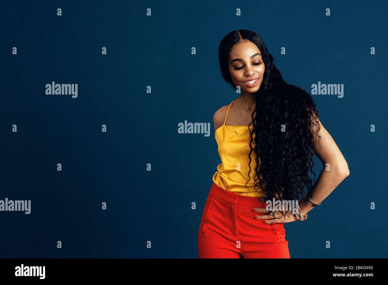 Portrait d'une jeune femme heureuse avec de très longs cheveux bouclés noirs souriants et regardant vers le bas, avec un bras à la taille Banque D'Images