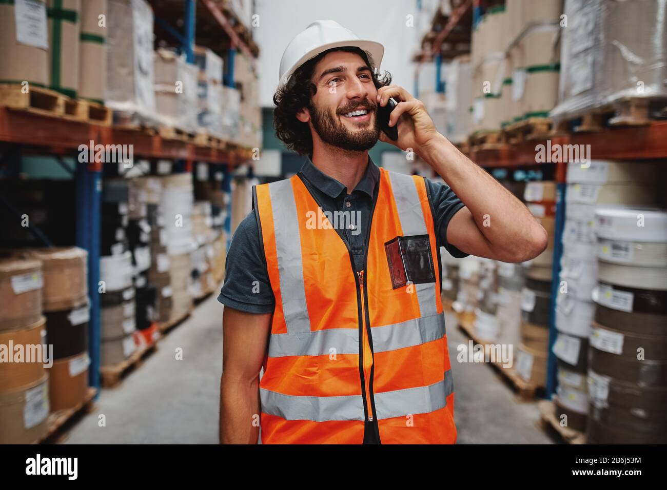 Un responsable d'entrepôt souriant lors d'une conversation sur un téléphone portable portant un casque blanc et un gilet de sécurité se tenant entre les rayons des marchandises et regardant à l'extérieur Banque D'Images
