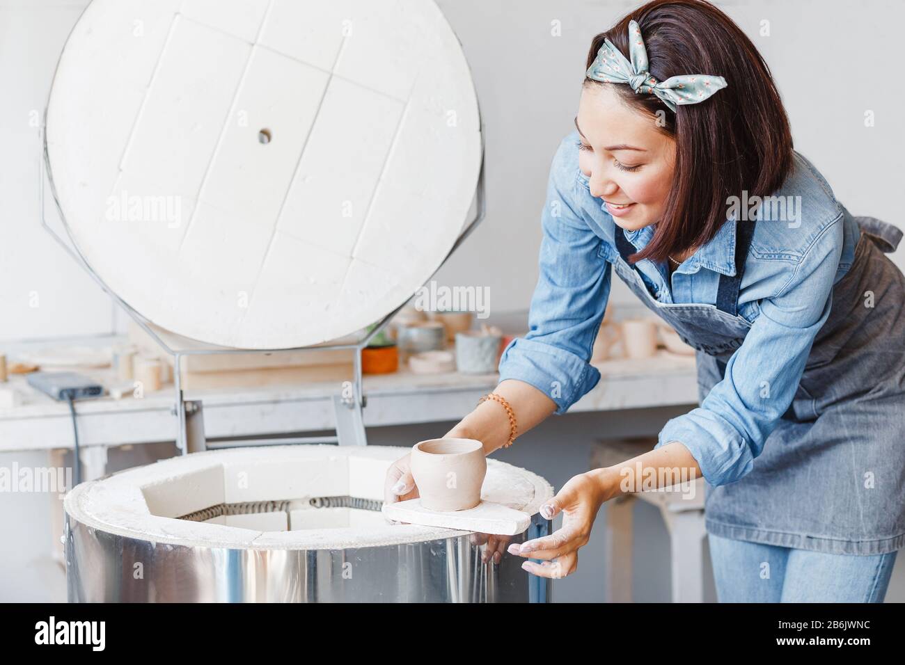 Jeune femme belle dans des vêtements de travail tenant un pot en argile dans ses mains se tenant près de la calcination et four à rôtir en atelier Banque D'Images