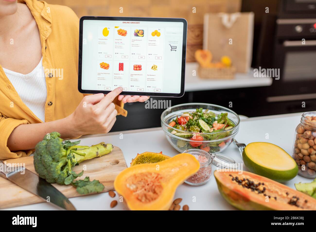Femme tenant une tablette numérique avec le marché de shopping en ligne lancé tout en se tenant sur la cuisine avec beaucoup de nourriture fraîche sur la table Banque D'Images